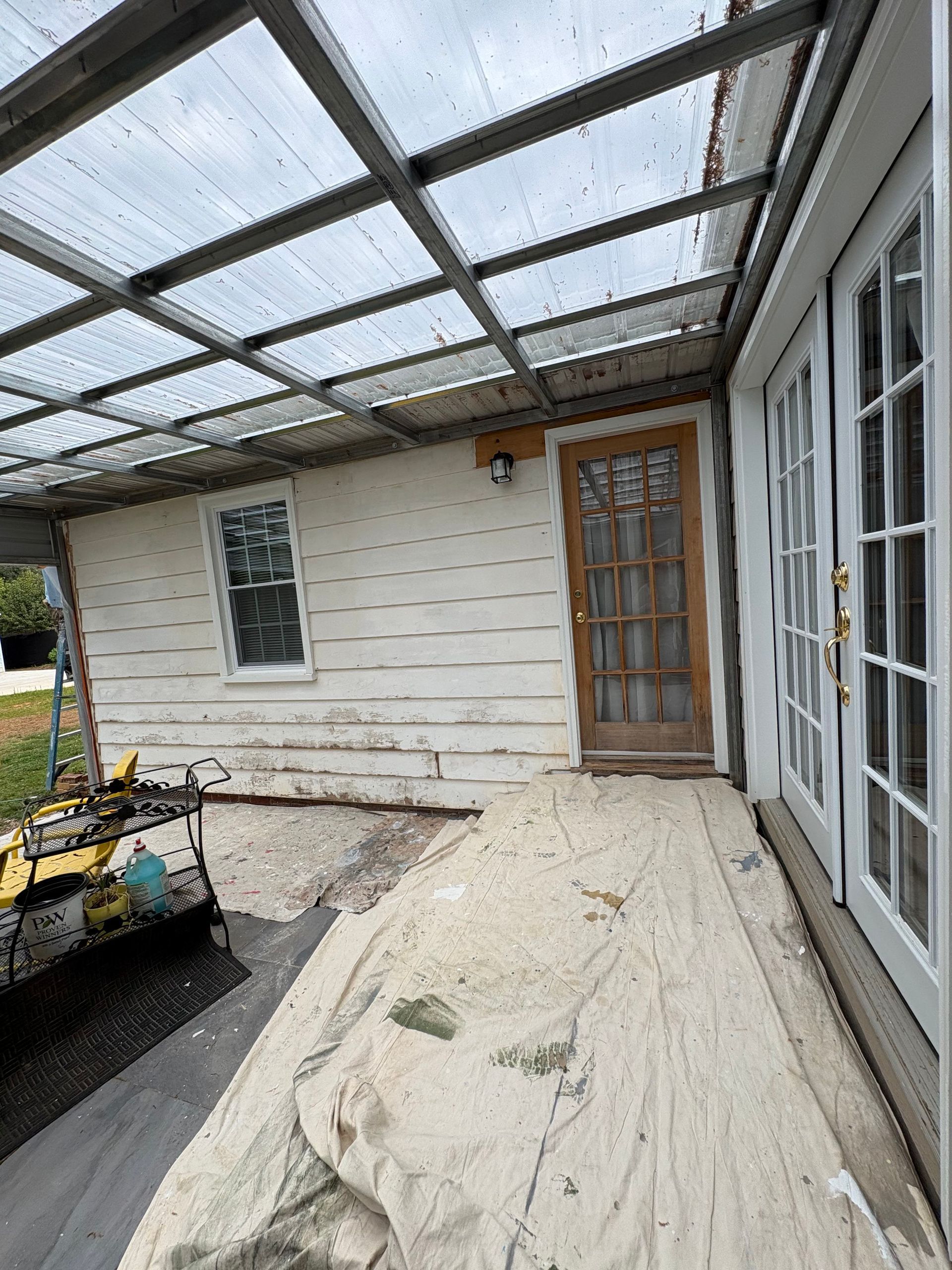 Covered porch with weathered wood siding, glass doors, and a wooden door under a transparent roof.