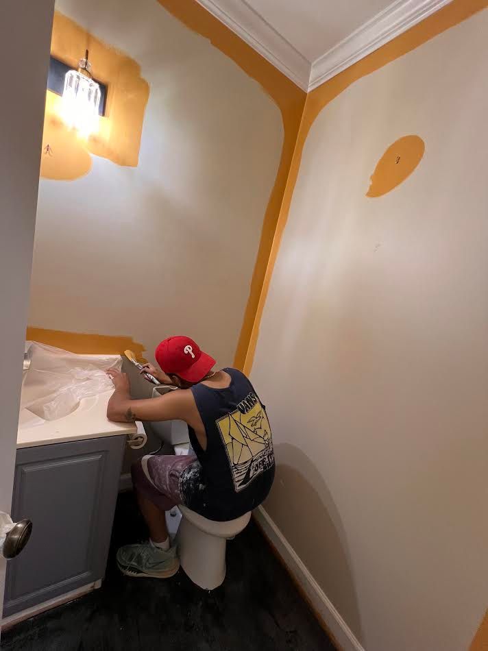 Person crouches beside a sink in a small bathroom, painting walls white with orange trim.