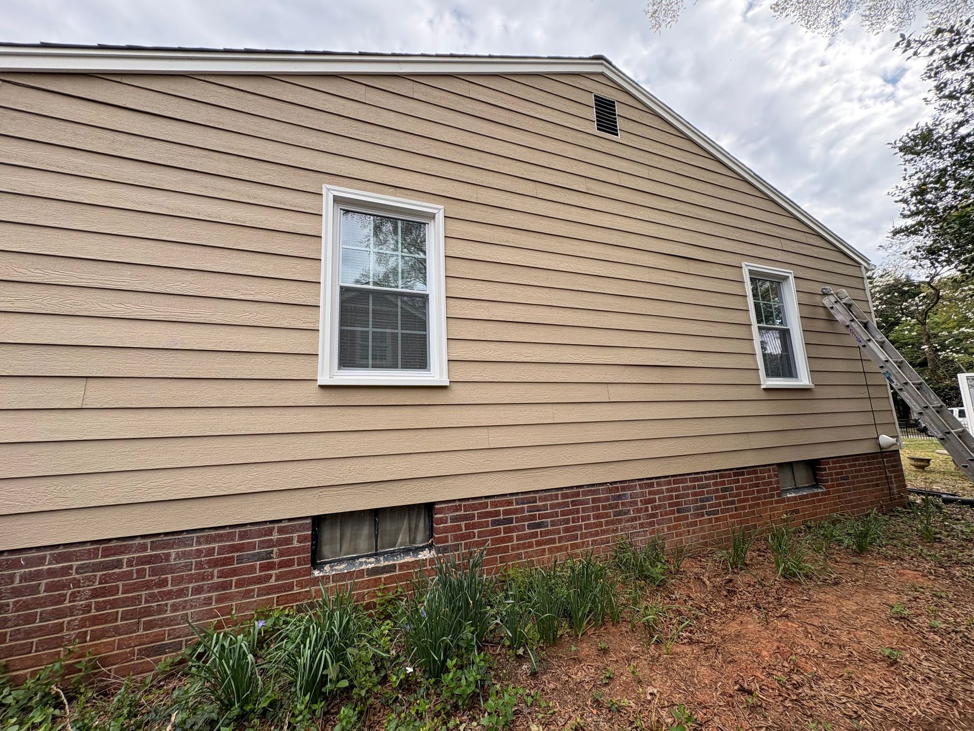 Side view of a beige house with white-trimmed windows, red brick foundation, and a small vent.