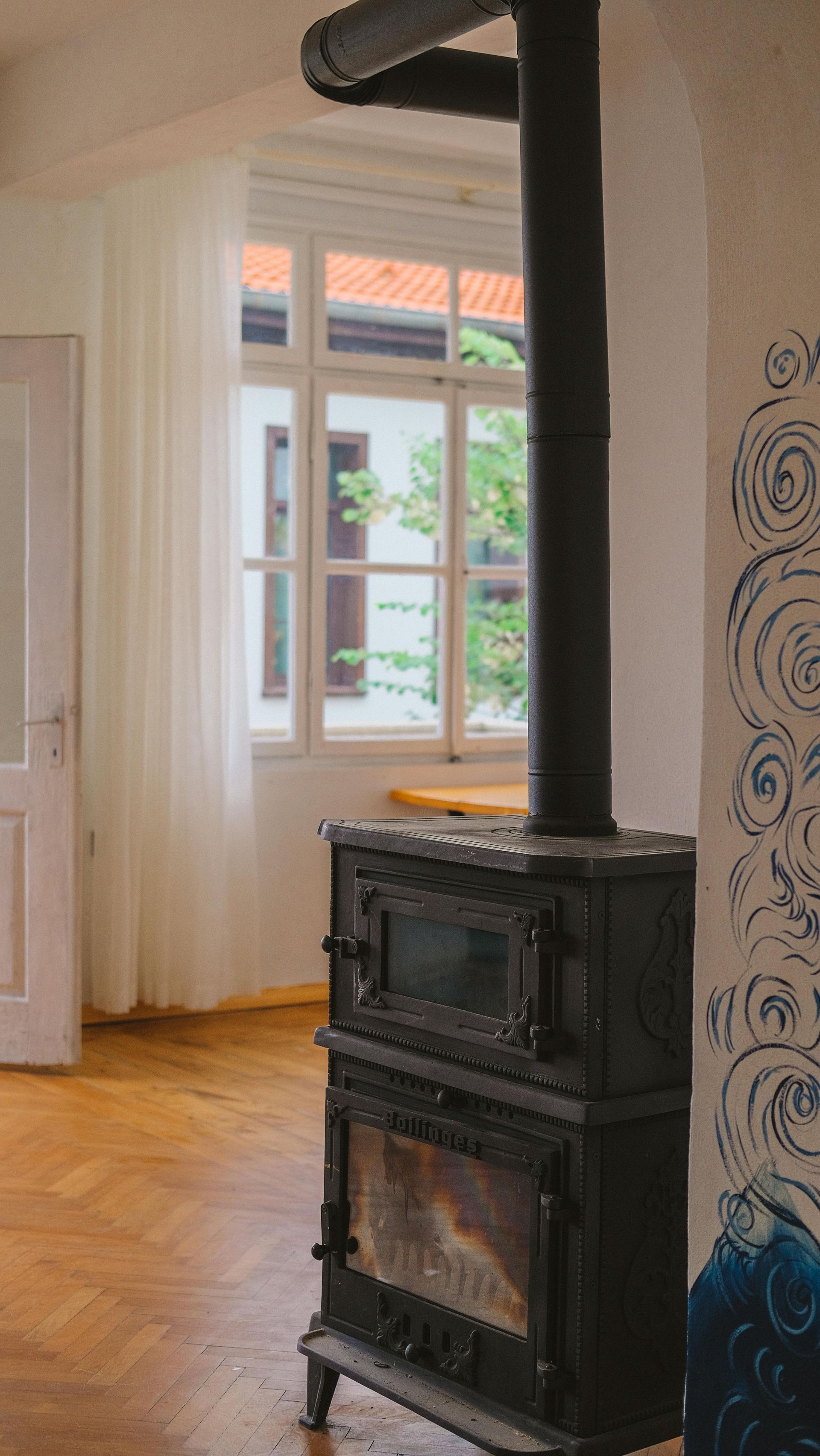 Black wood-burning stove with chimney in a room with a window and a sheer white curtain.