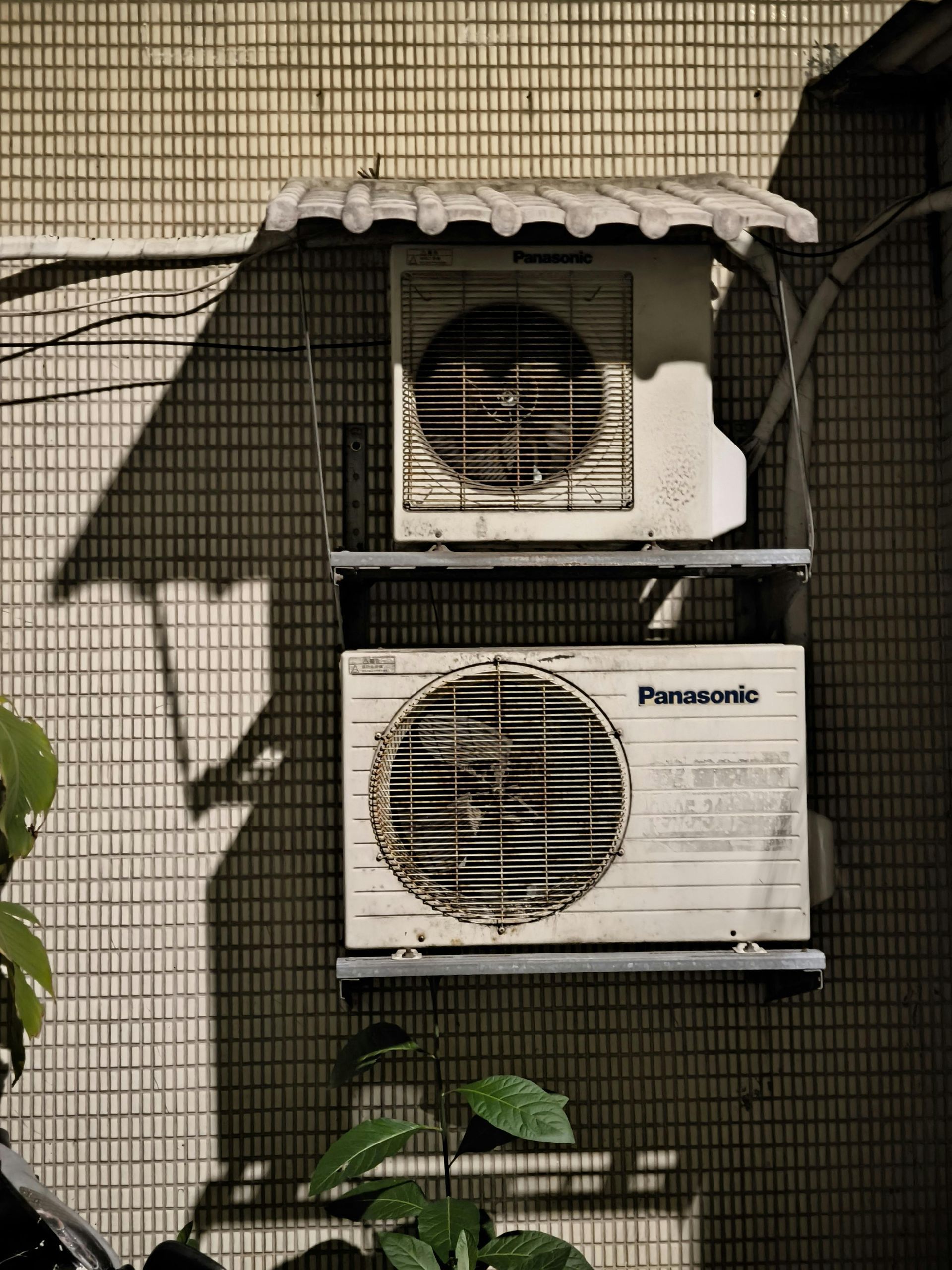 Two air conditioning units, Panasonic brand, stacked on a tiled wall, beneath a small roof, with shadows and plants.