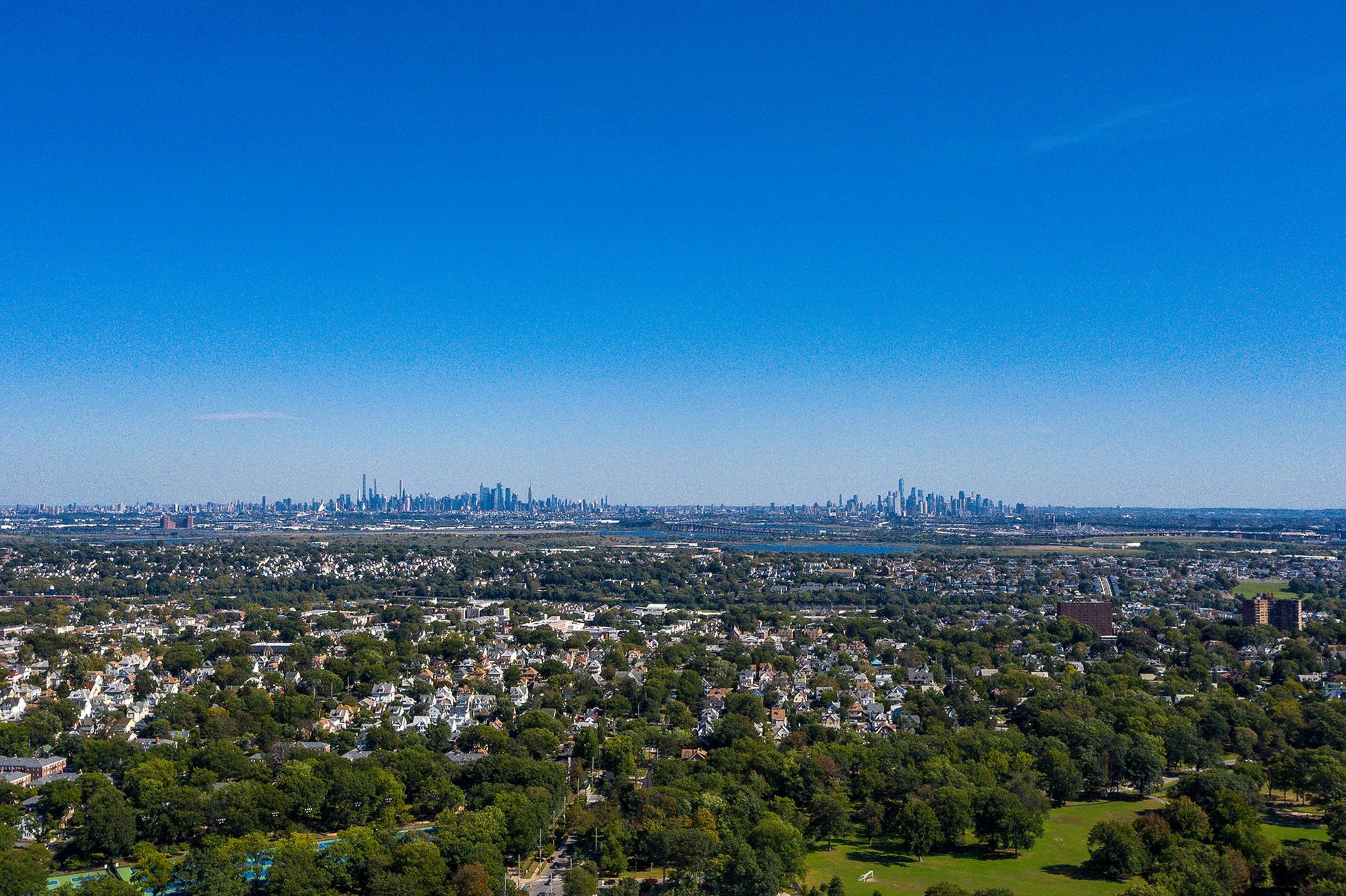 Aerial view of a city with a skyline in the distance, lush green trees and houses dominate the foreground under a blue sky.