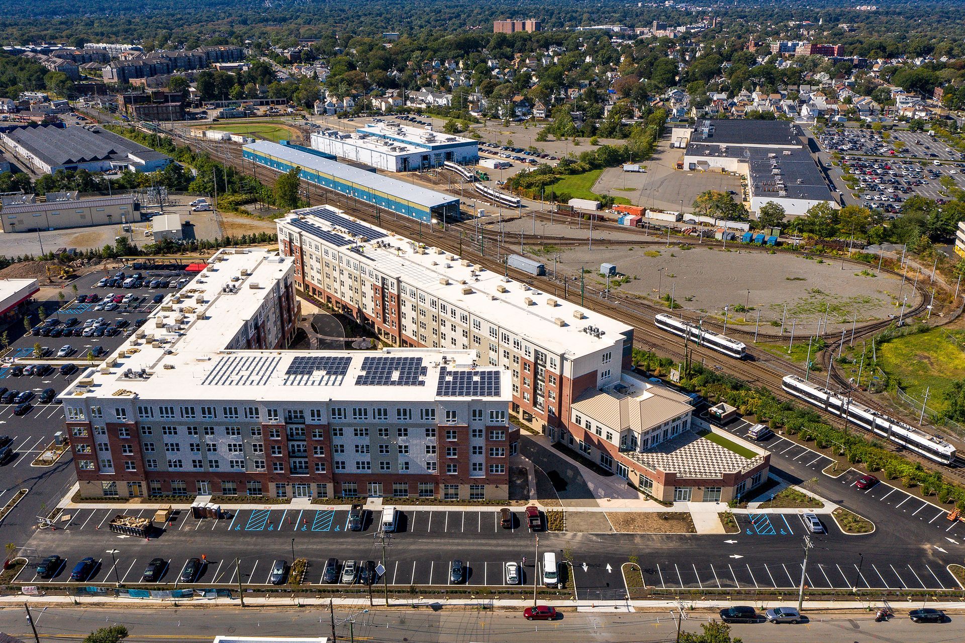 Aerial view of a multi-story apartment building with a parking lot, near train tracks and a station.