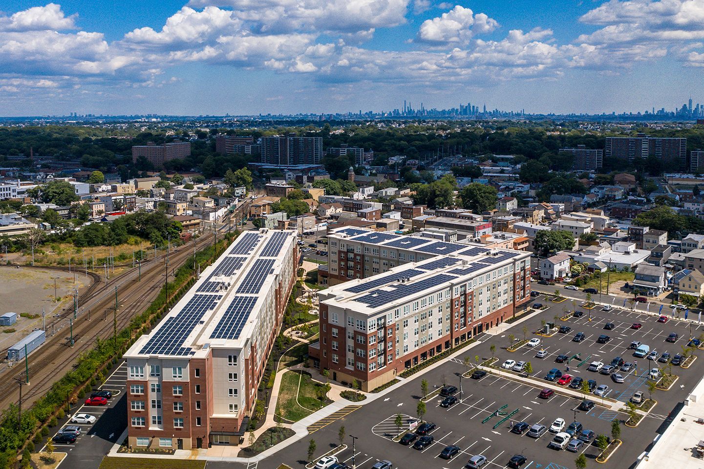 Aerial view of apartment buildings with solar panels, surrounded by parking and cityscape.