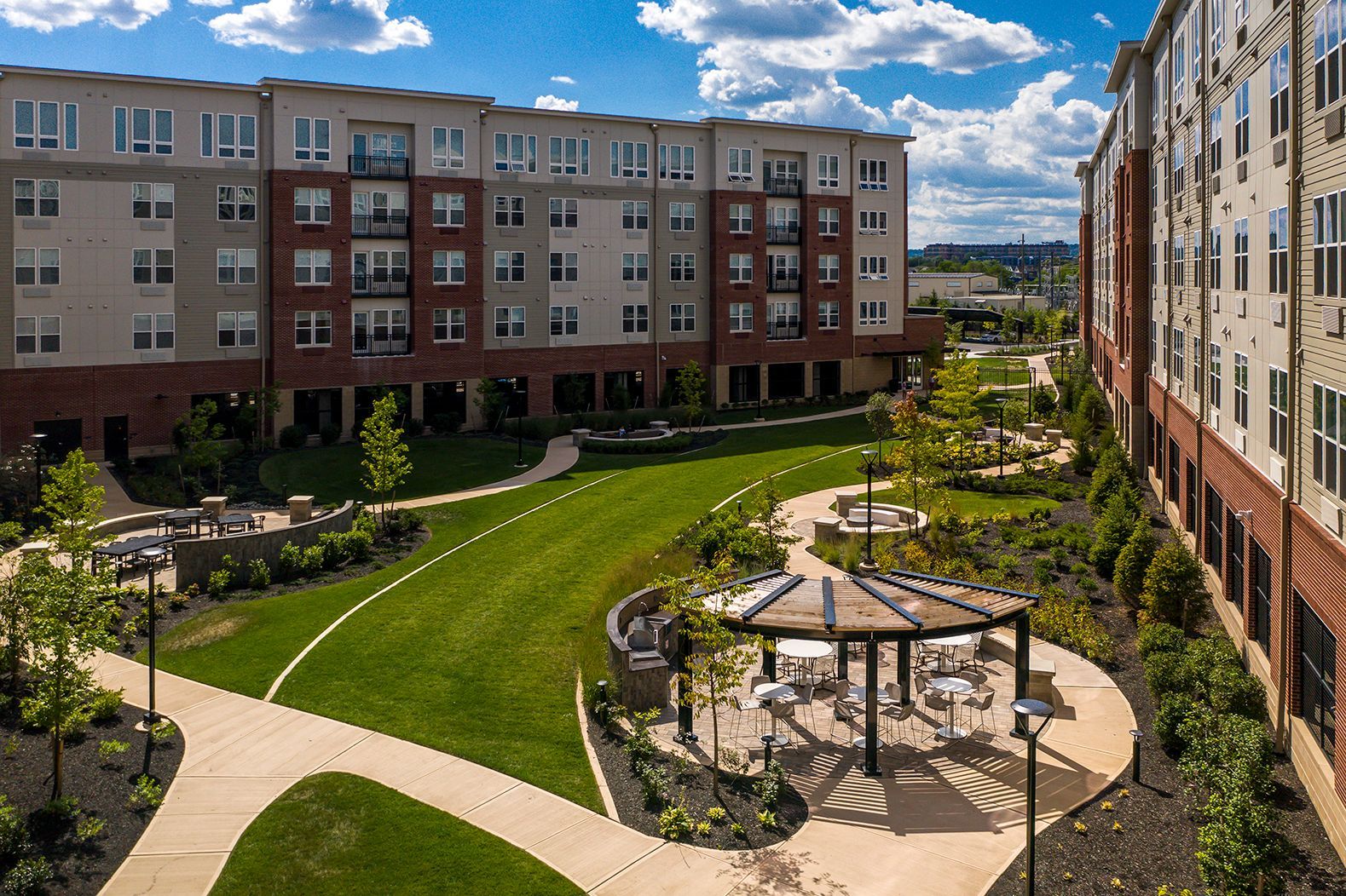 Exterior of apartment buildings with a courtyard featuring a lawn, gazebo, and landscaping under a sunny sky.