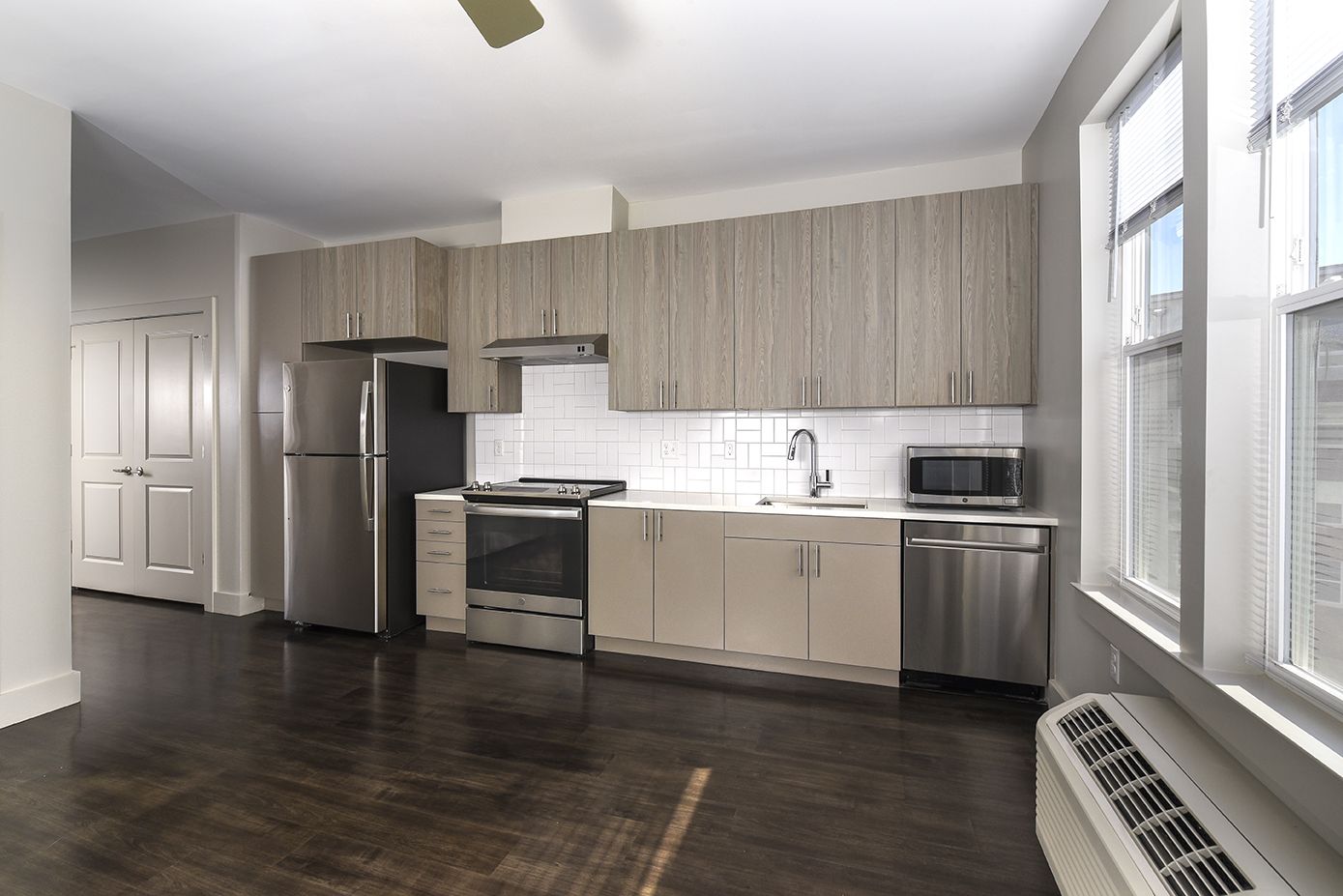 Modern kitchen with stainless steel appliances, light wood cabinets, dark wood floors, and two windows.