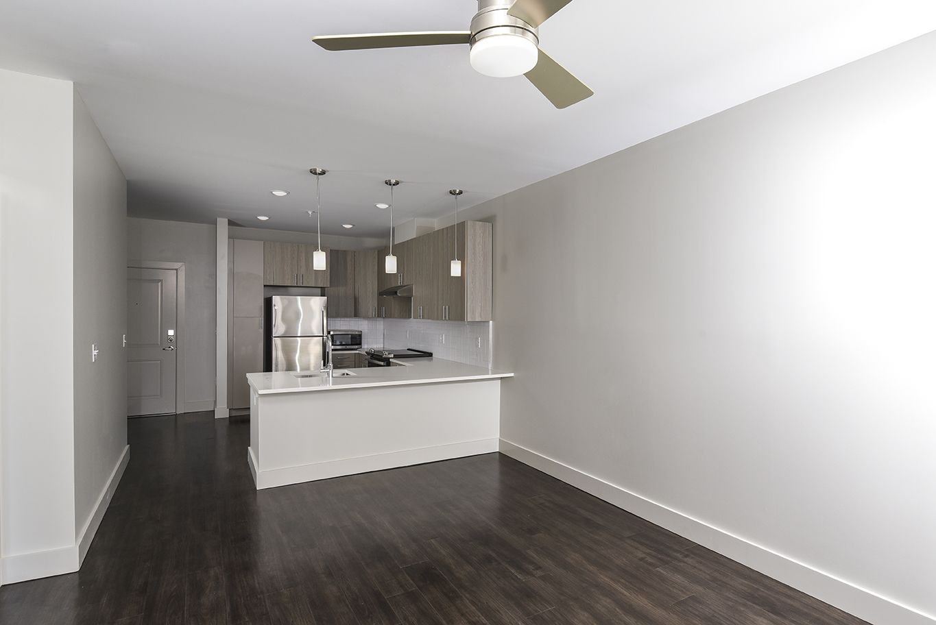 Empty modern apartment with dark wood floors, white walls, and kitchen area.