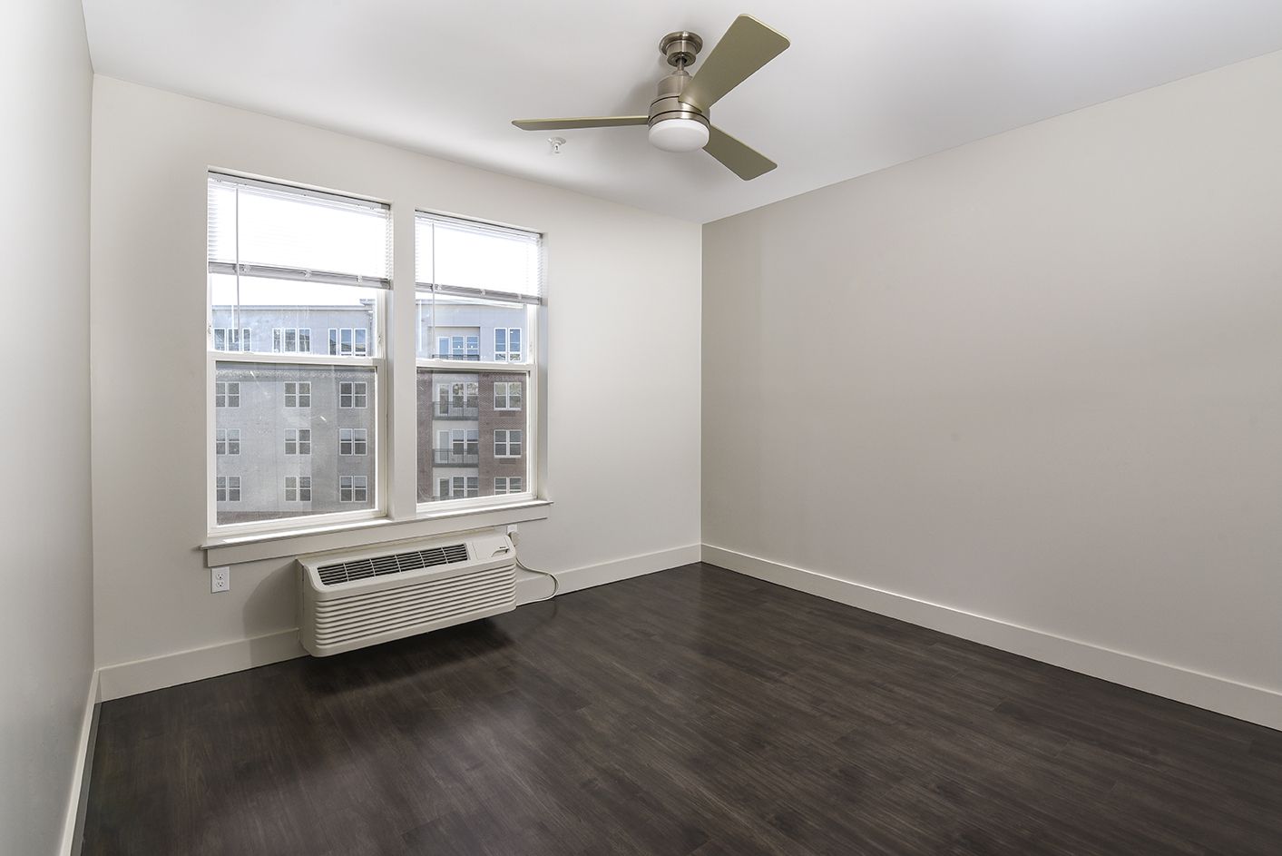 Empty bedroom with dark wood floor, large window, ceiling fan, and beige walls.