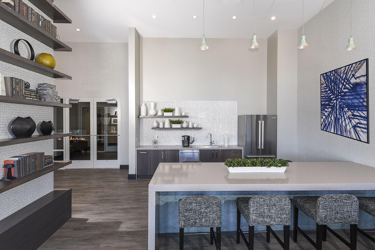 Communal lounge with kitchen, shelves, long counter, patterned bar stools, and abstract art on the wall.