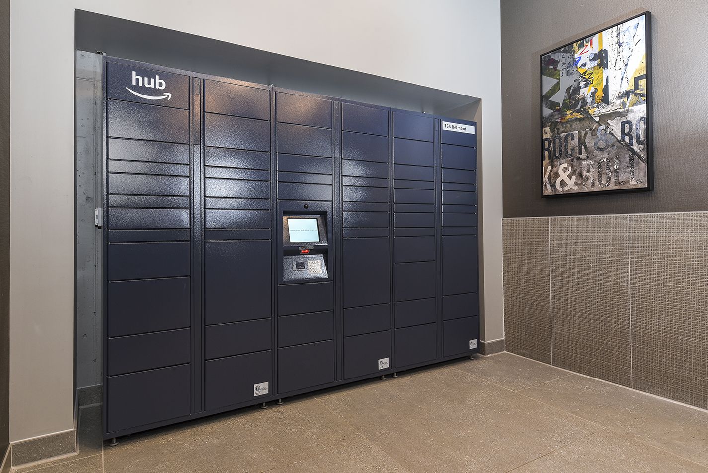 Dark blue package lockers with a central touchscreen in a building hallway.