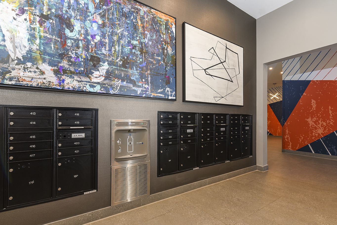 Mailboxes and water fountain in an apartment hallway, artwork on the wall, and entrance.