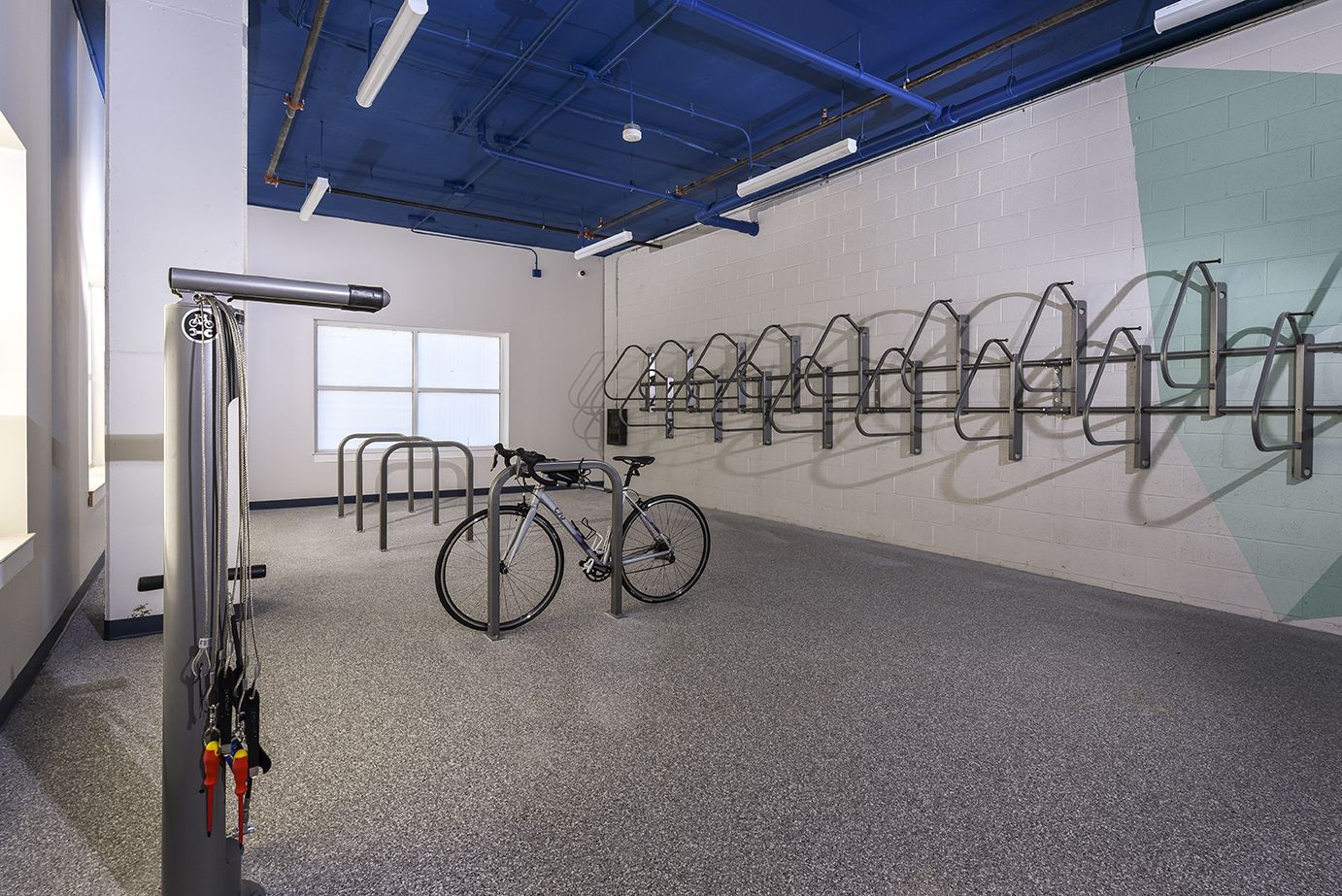 Bike storage room with multiple racks, one bicycle parked, and a work station. Blue ceiling and patterned floor.