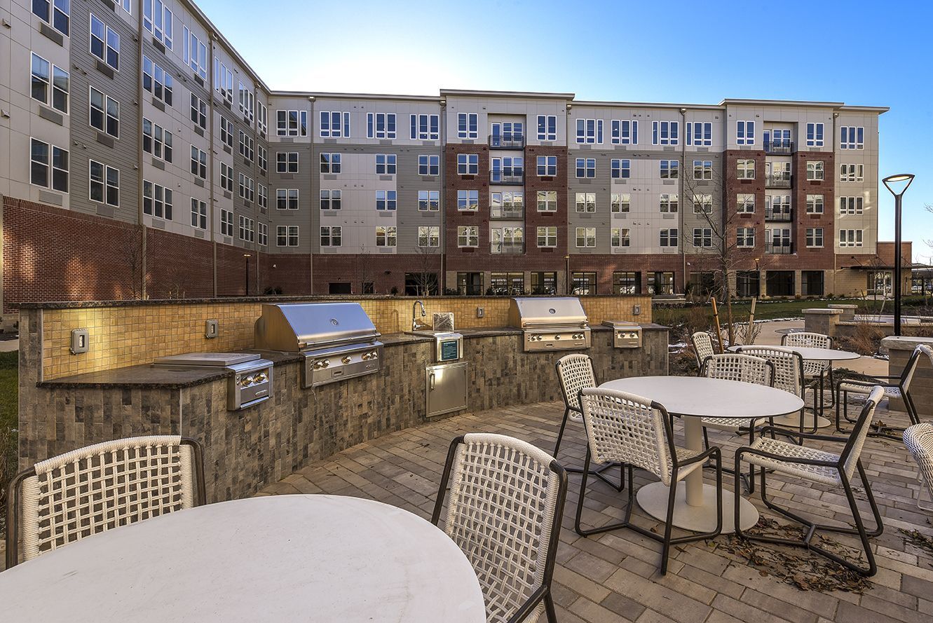 Outdoor grilling area with built-in grills, tables, and chairs in front of a multi-story apartment building.