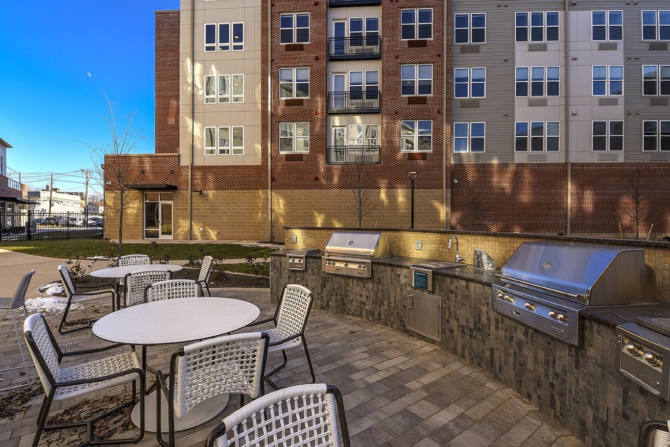 Outdoor grilling area with tables, chairs, and several stainless steel grills against a multi-story brick building.