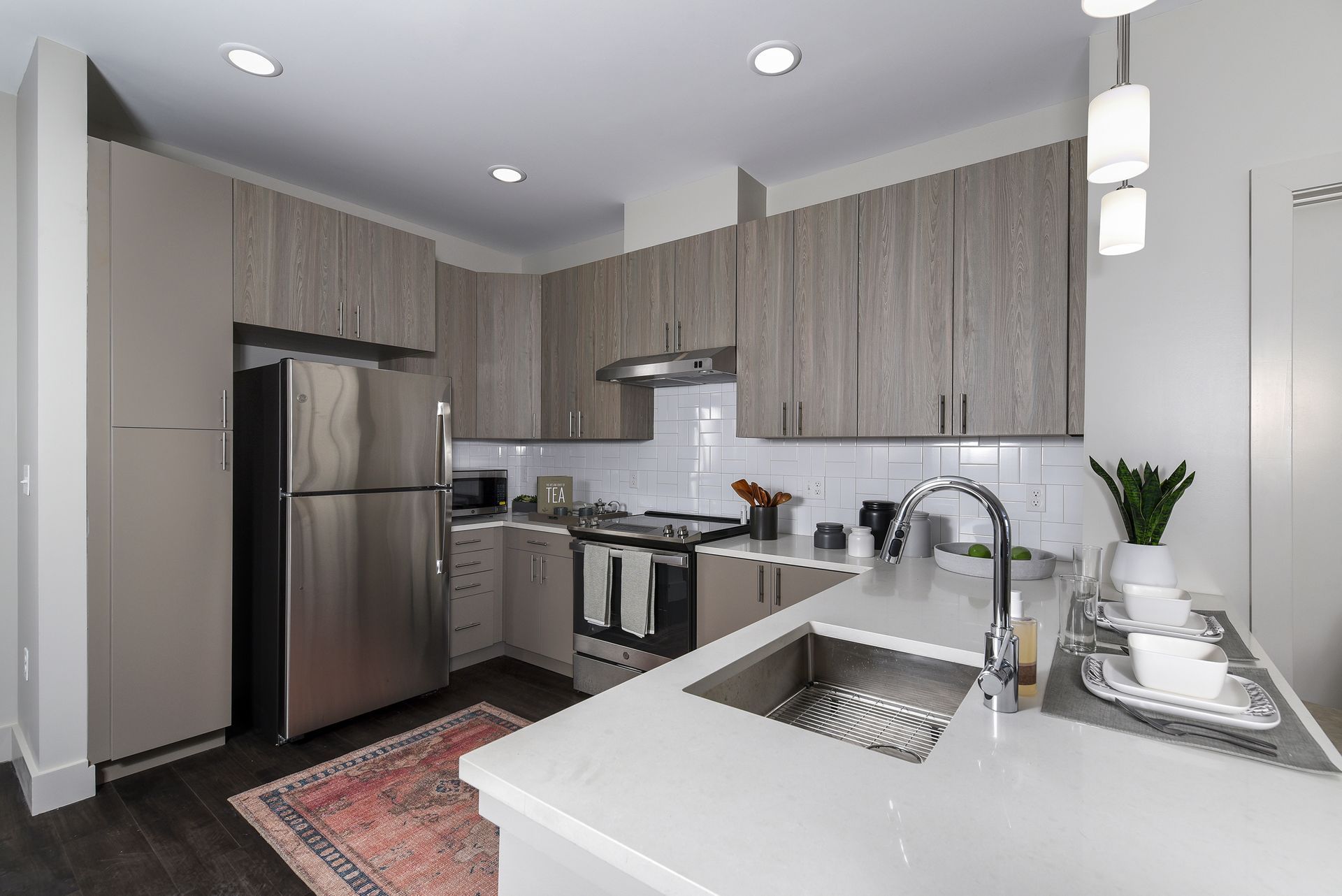 Modern kitchen with stainless steel appliances, light wood cabinets, white countertops, and a patterned rug.