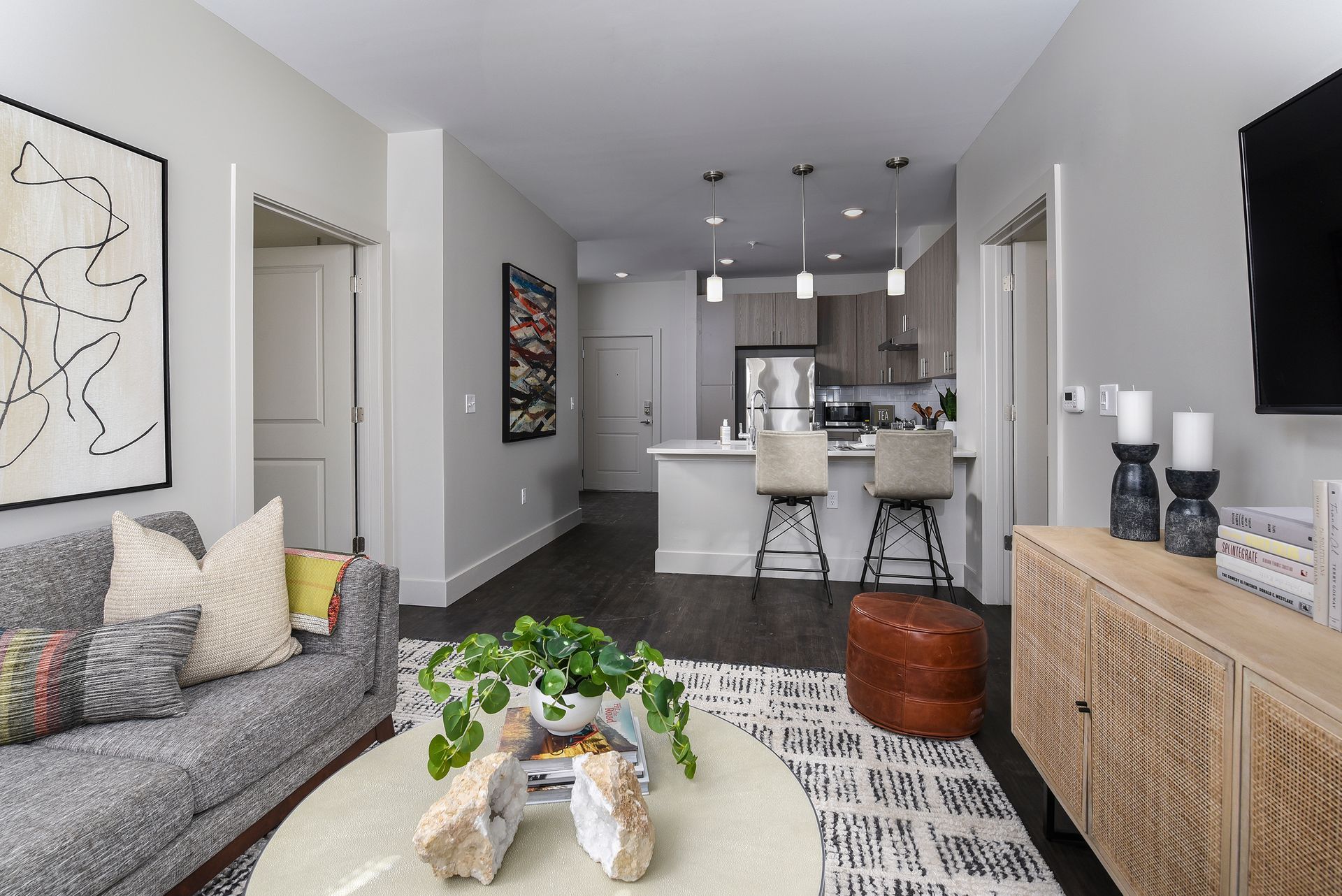 Modern living room with kitchen in the background. Gray sofa, wooden cabinet, and abstract art.