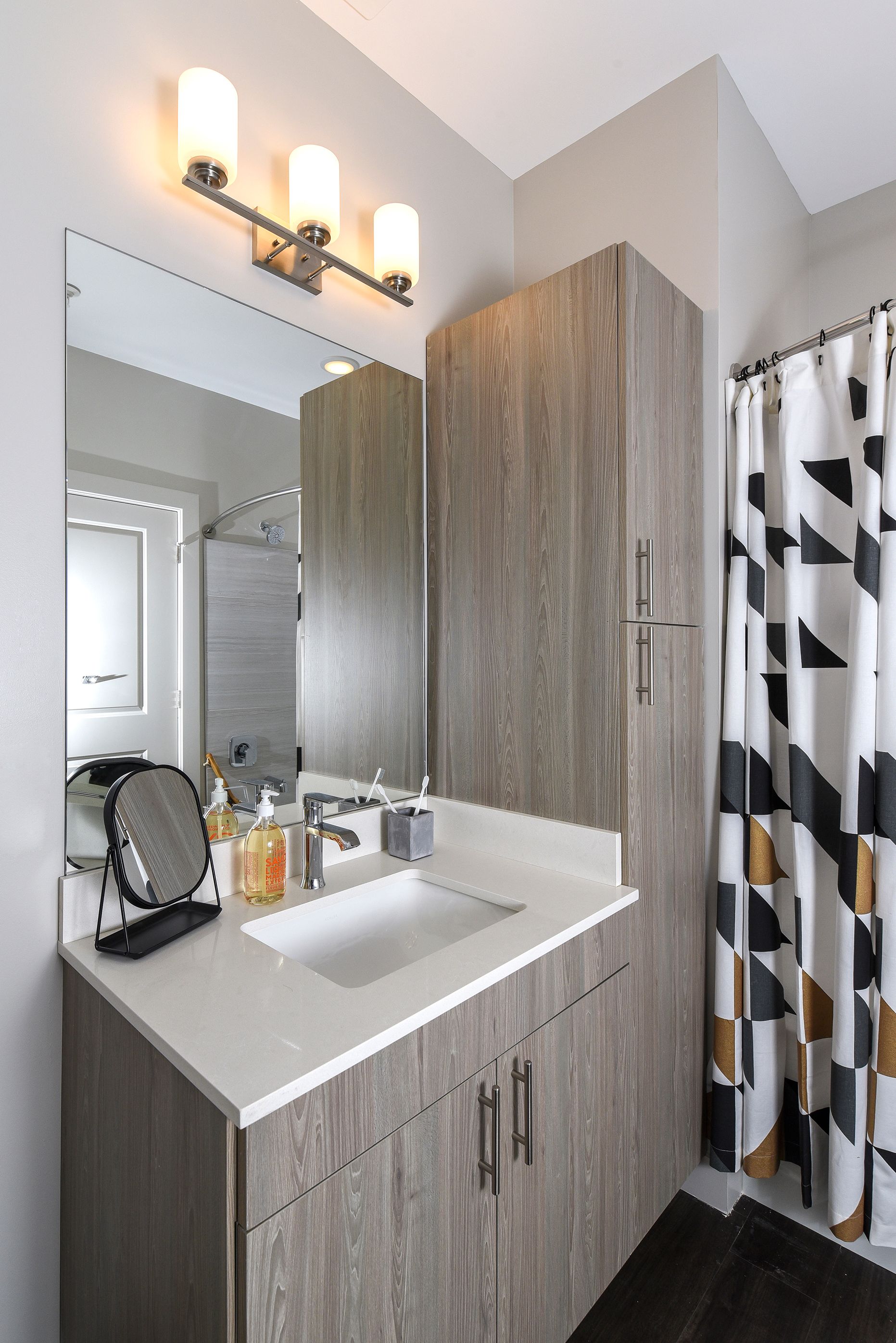 Bathroom with wood-look cabinets, white countertop, large mirror, and a geometric pattern shower curtain.