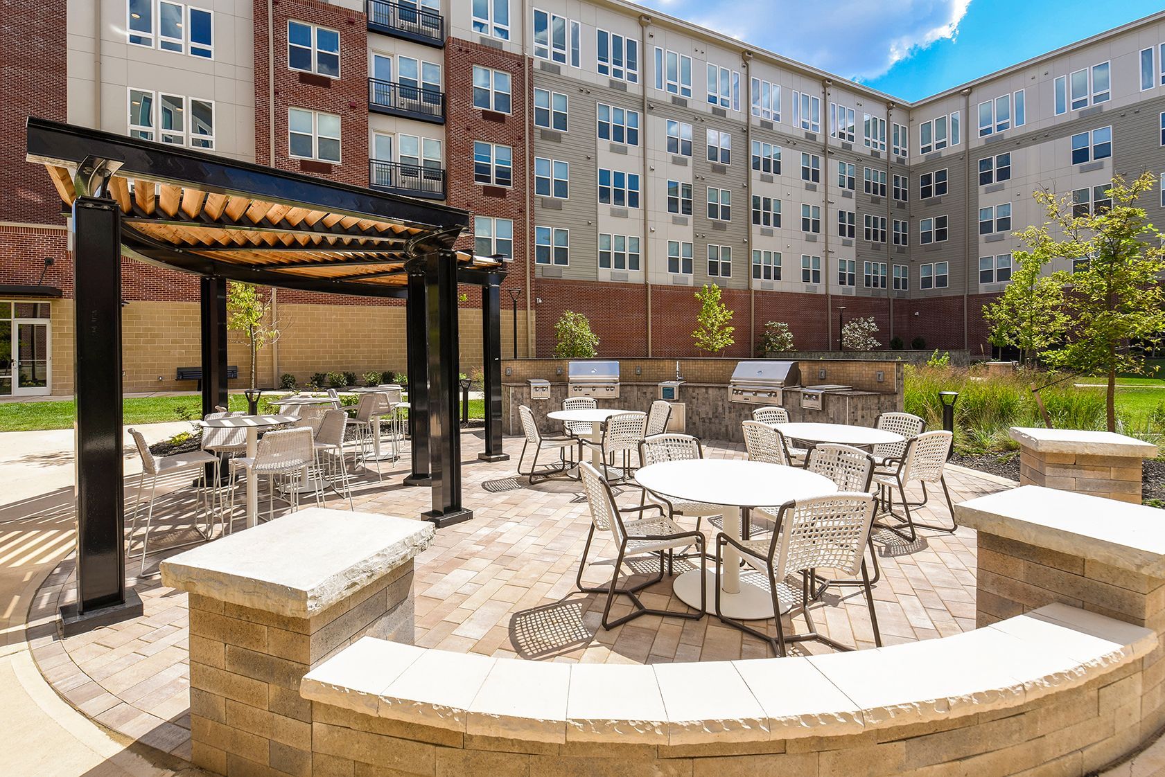 Courtyard with tables, chairs, pergola, and grills. Brick and light-colored buildings in background. Sunny day.
