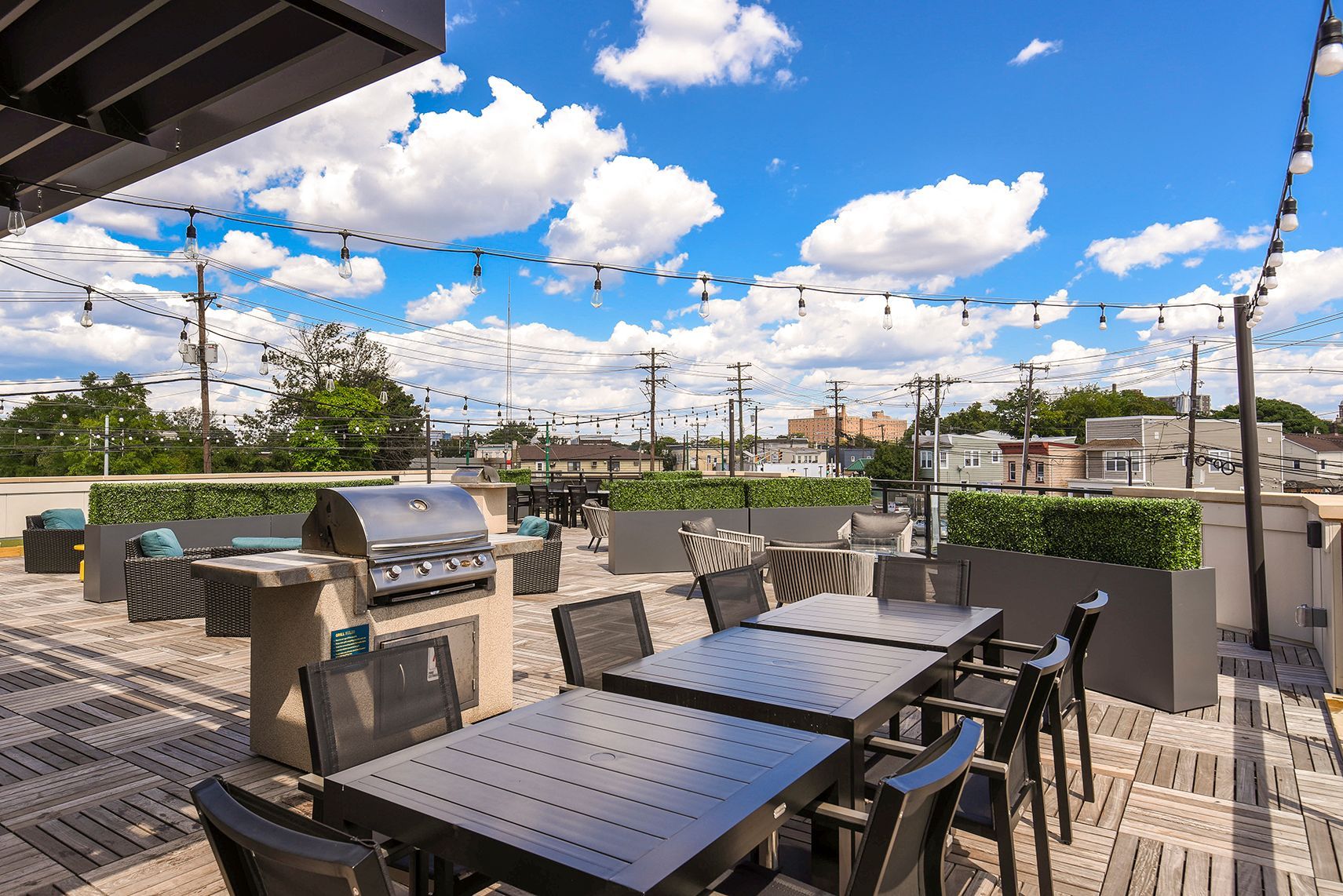 Rooftop patio with grill, seating, and string lights against a blue sky with fluffy clouds.
