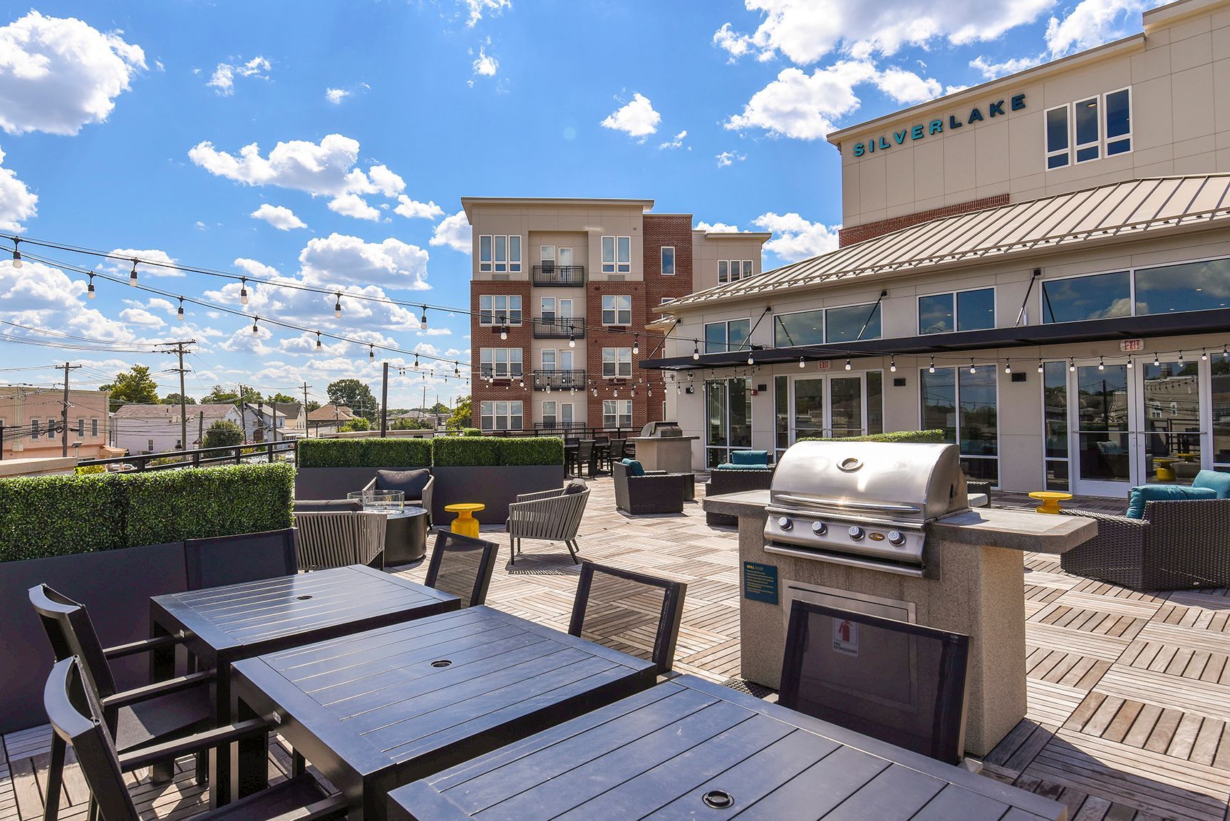 Rooftop patio with grilling area, tables, and lounge seating, under a sunny, blue sky with fluffy clouds.