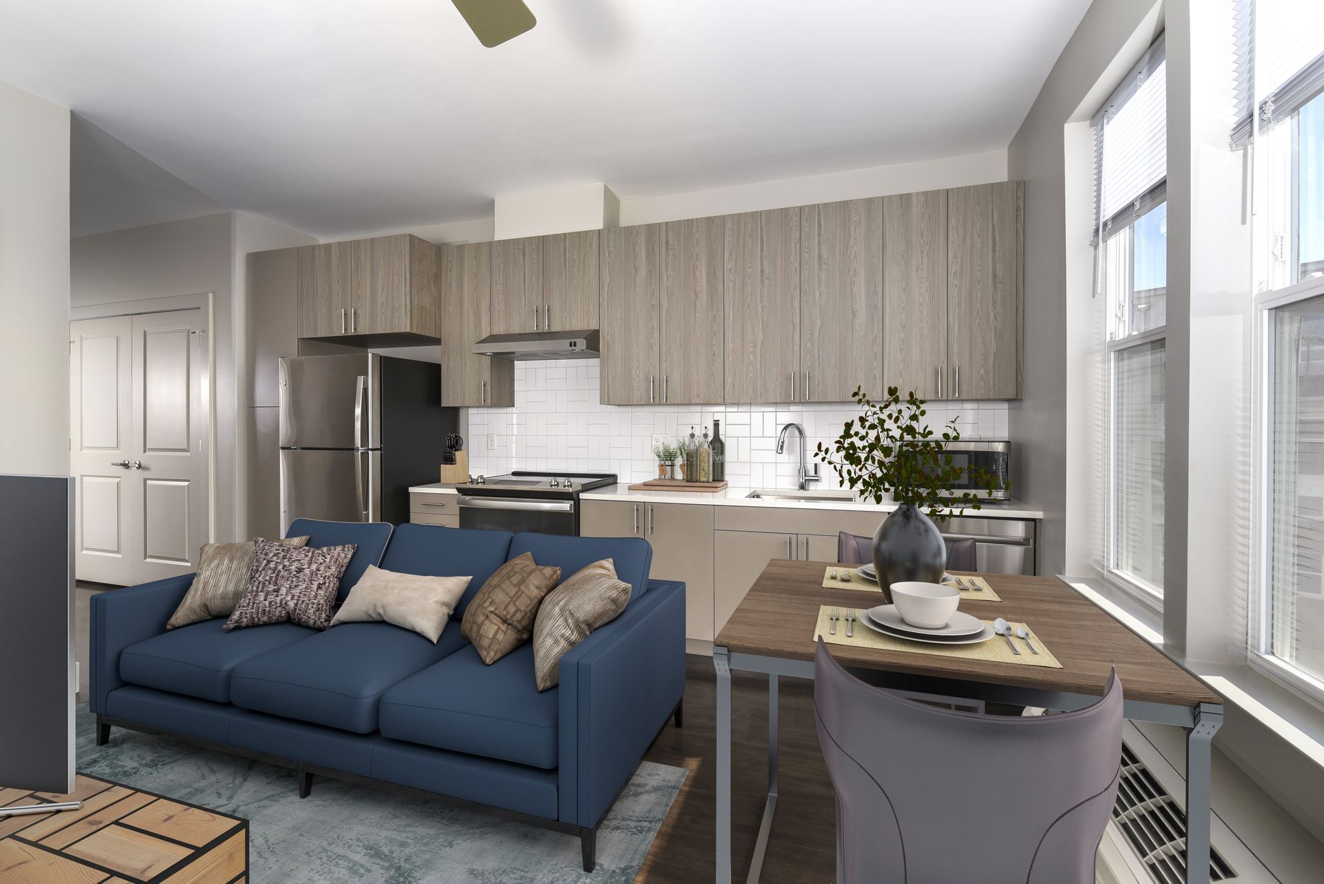 Modern apartment interior with blue couch, wooden cabinets, and dining table near windows.