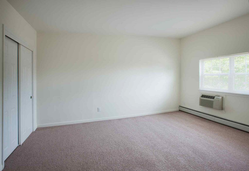 A carpeted bedroom with a window, an air conditioning unit and a closet
