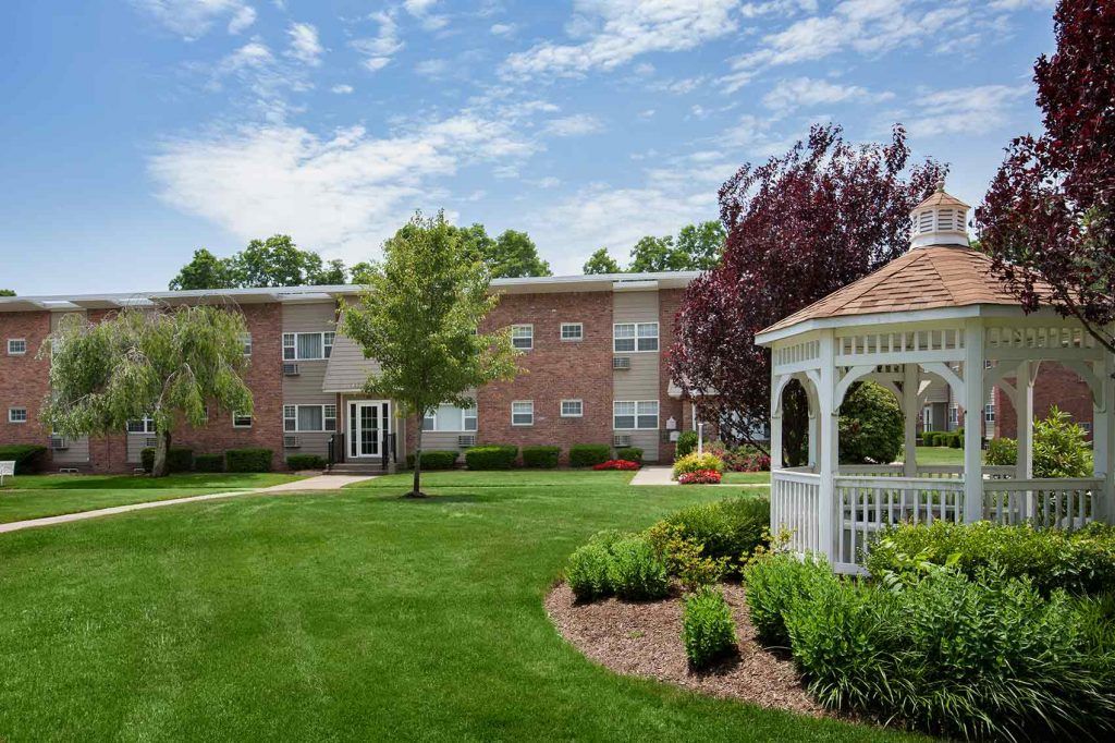 Gazebo with landscaping and grass surrounding it. Two story, brick apartment building in background