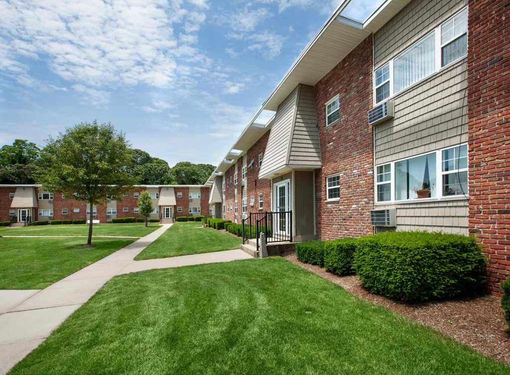 Brick front apartment building with bushes, grass, and trees in front, with sidewalks connecting them