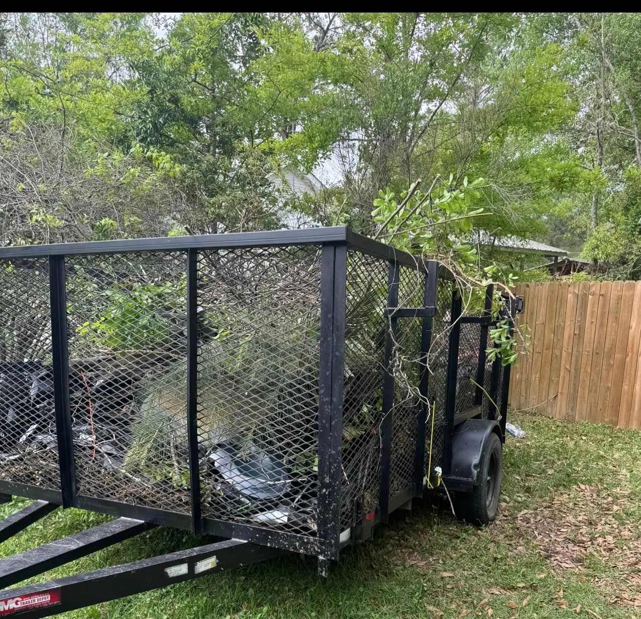 A trailer filled with junk is parked in the grass next to a wooden fence.