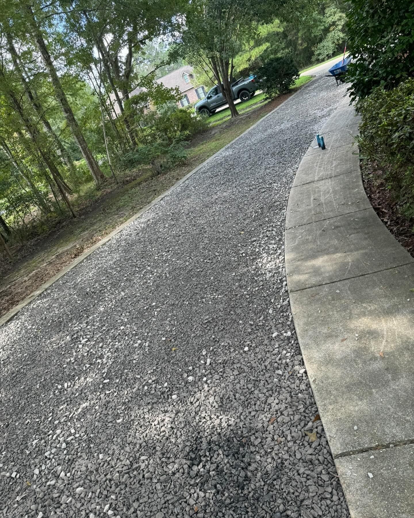 A concrete walkway leading to a gravel driveway surrounded by trees.