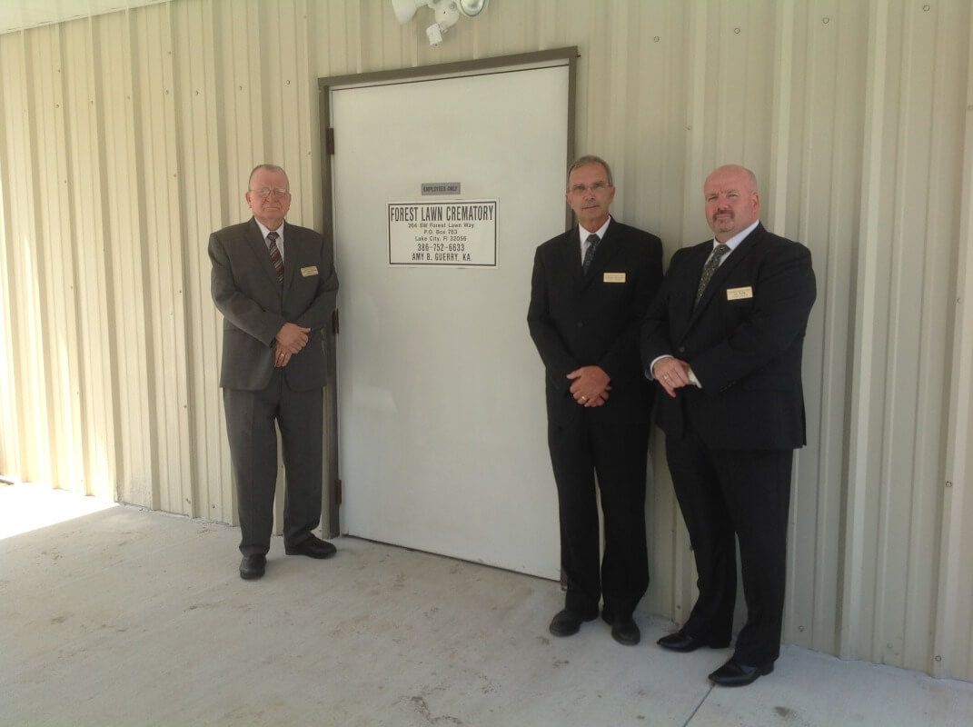 Three men in suits stand in front of a white door with a sign on it. They are in front of a metal building.