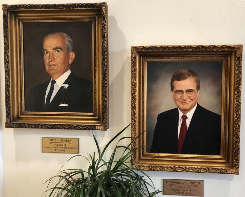 Two framed portraits of men in suits on a wall, with a plant below.
