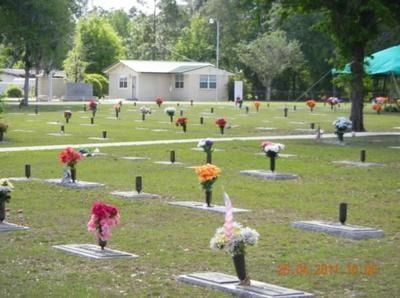 Cemetery with headstones and vases of flowers; small building in the background. Green grass and trees surround.