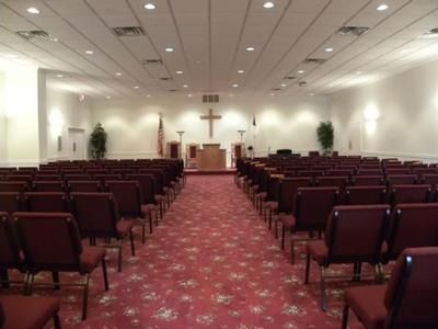 Interior of a church sanctuary with rows of chairs facing a cross and podium. Red carpet and white walls.