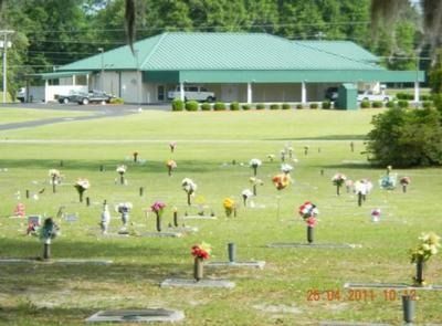 Cemetery with headstones and flowers in foreground, building with green roof in background.