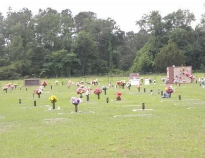 Cemetery with rows of headstones, floral arrangements, and green grass against a backdrop of trees.