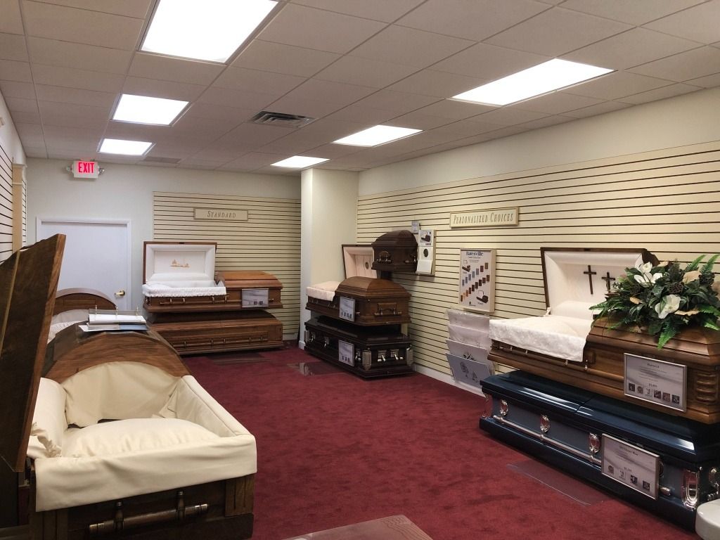 Interior of a funeral home displaying several open coffins on red carpet; neutral lighting.