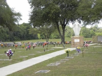 Cemetery with headstones and a statue of a person on a pedestal; a pathway leads through the green space.