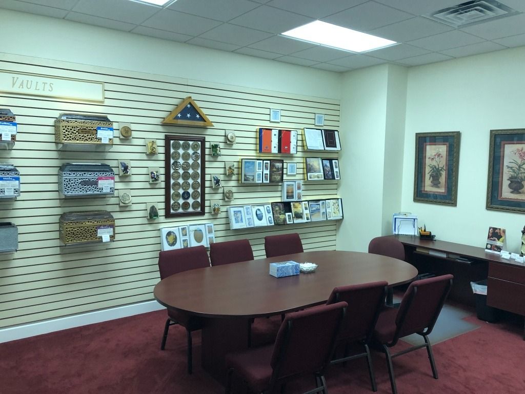 A room in a funeral home; oval table, chairs, displays of urns, flags, and framed photos.