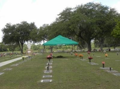 A cemetery with a green tent set up for a service, flowers at grave markers, and trees.