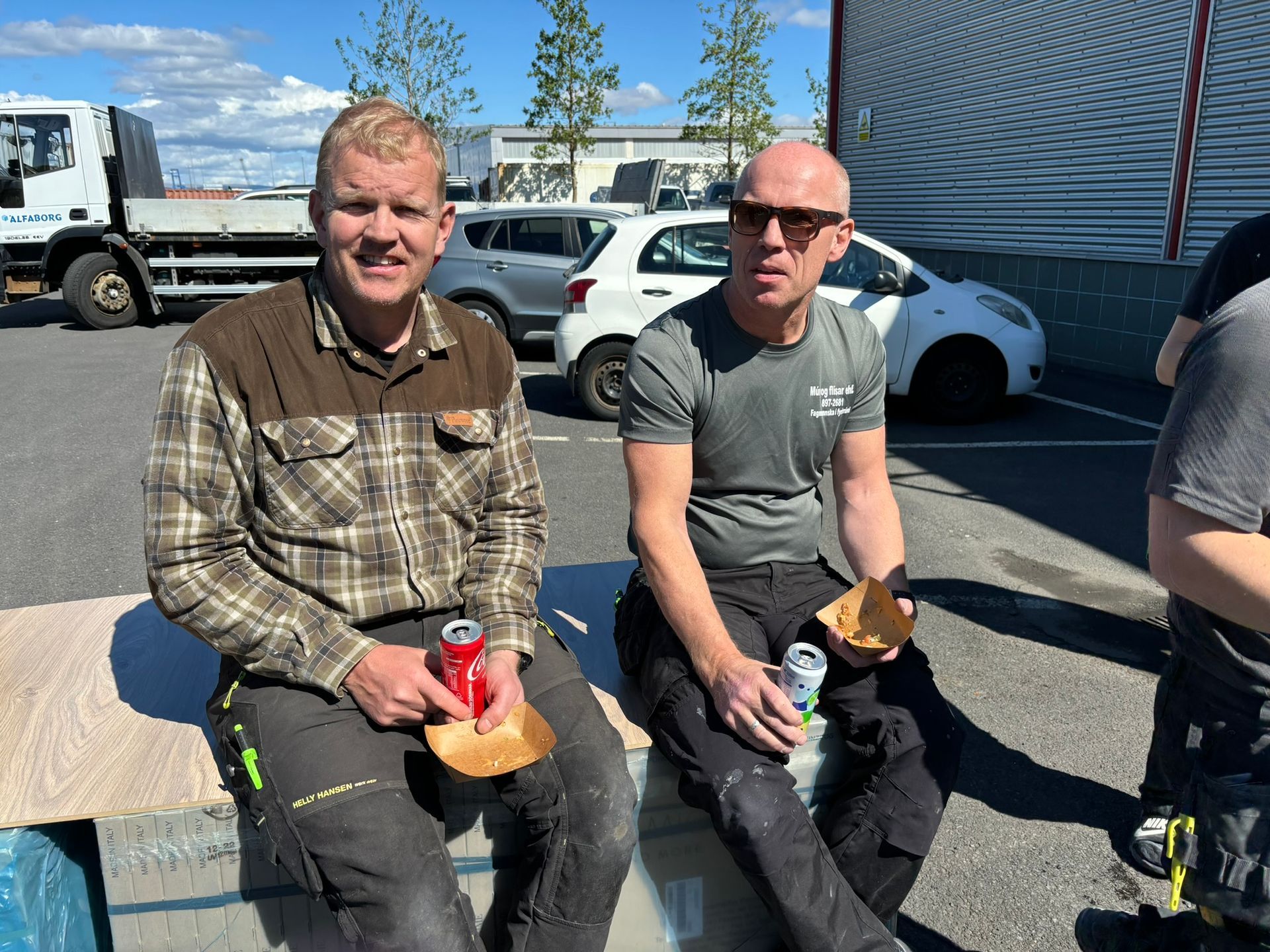 Tveir menn sitja á bekk á bílastæði að borða mat og drekka gos.