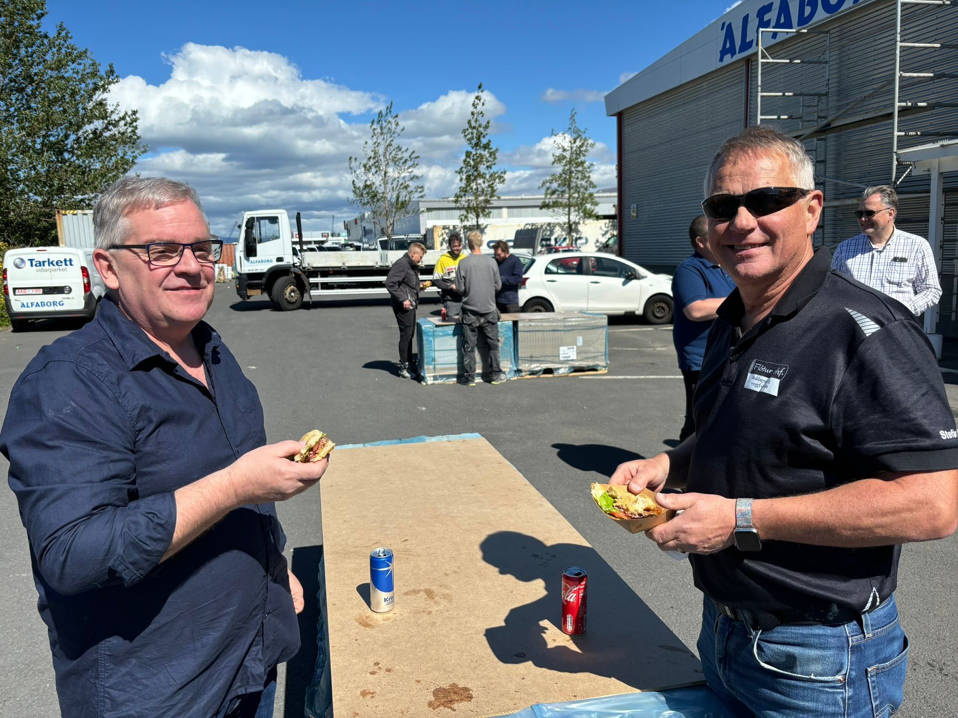 Tveir menn standa við hlið hvors annars á bílastæði og borða samlokur.