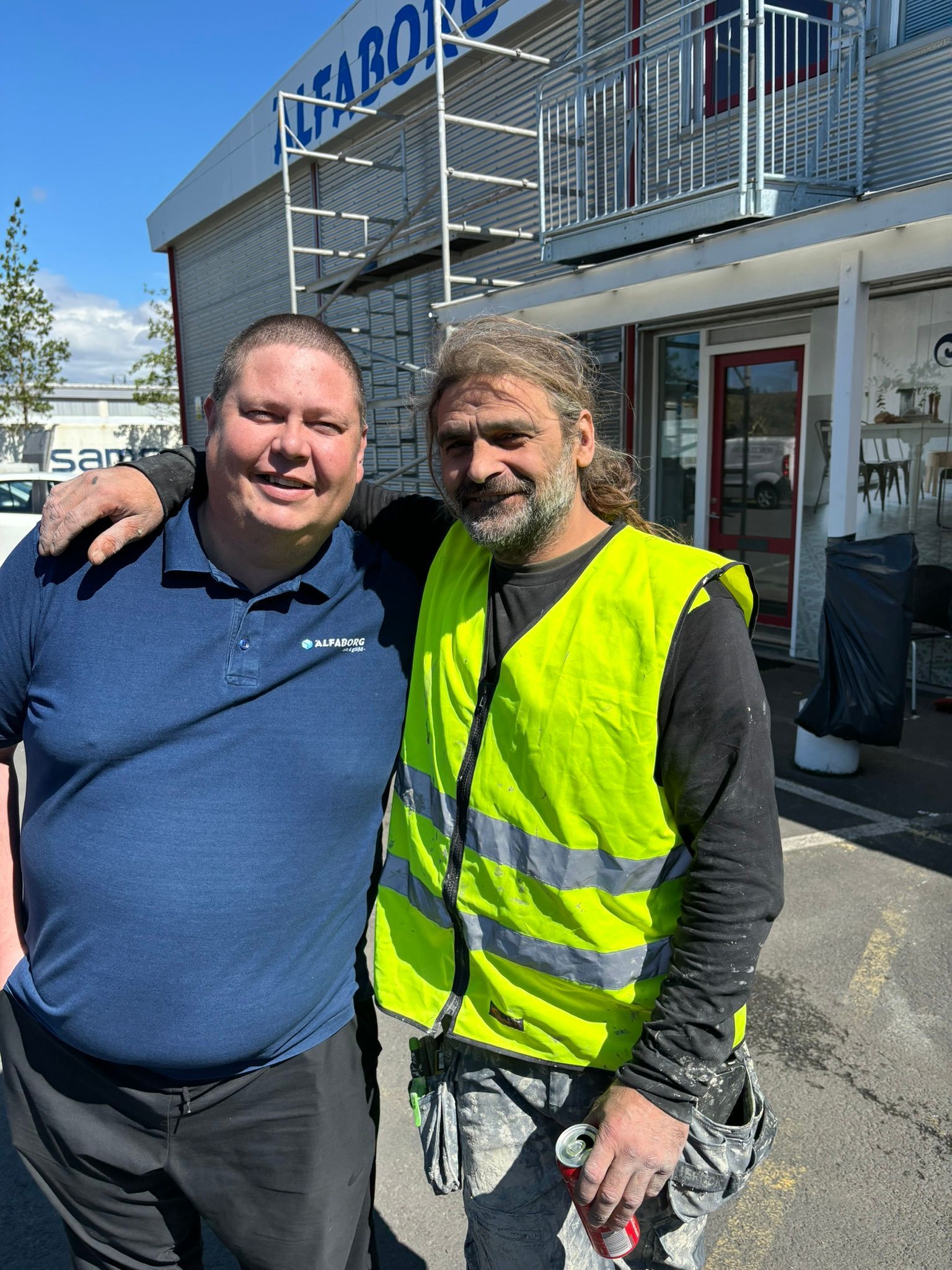 Tveir menn sitja fyrir á mynd fyrir framan byggingu. annar mannanna er í gulu vesti.