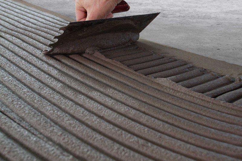 Maður er að dreifa lími á flísalagt gólf með spaða.