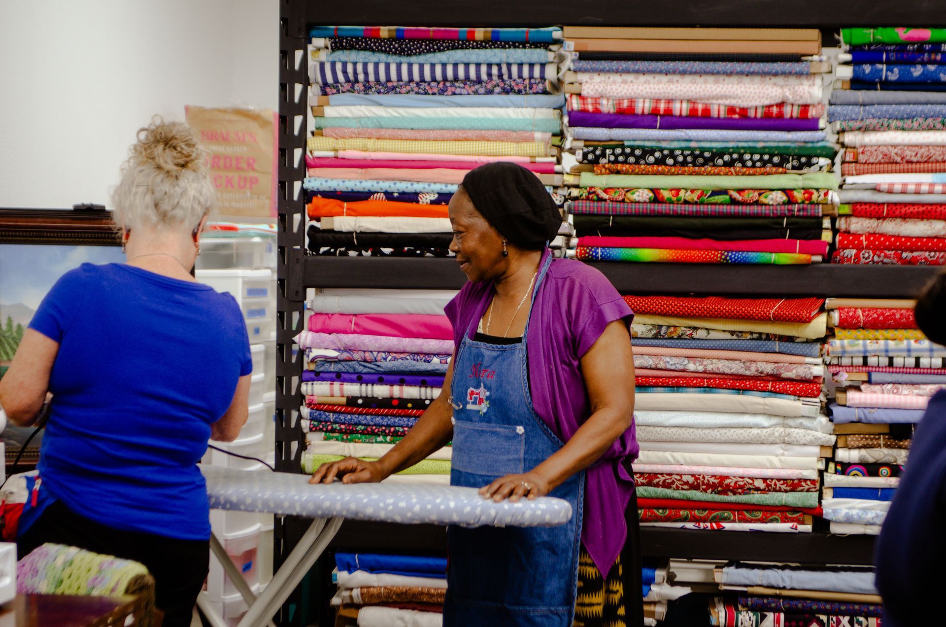 A woman is standing at an ironing board in front of a shelf of fabric.
