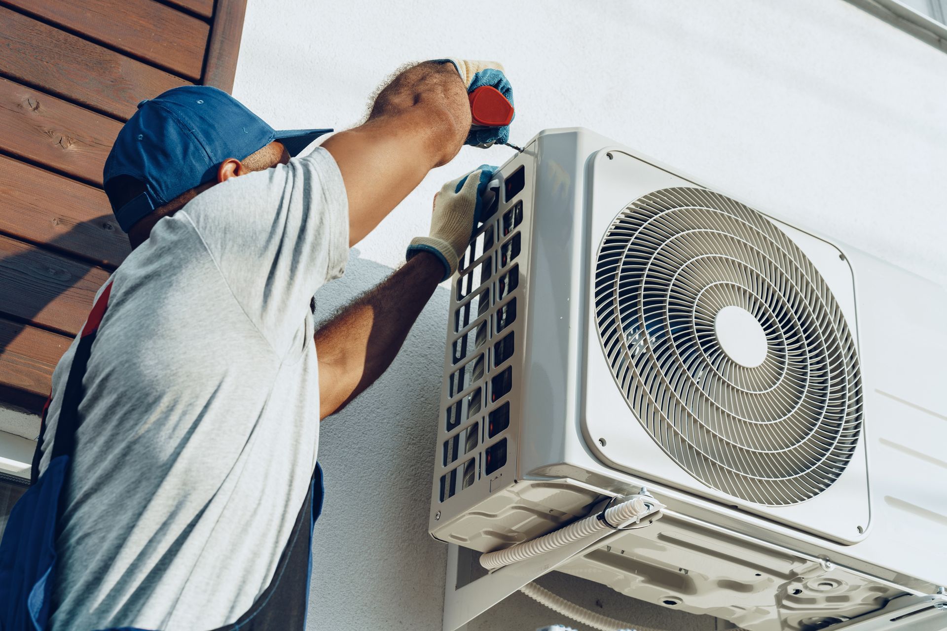 A technician in a blue cap and work clothes repairs a white outdoor air conditioning unit mounted on a wall.