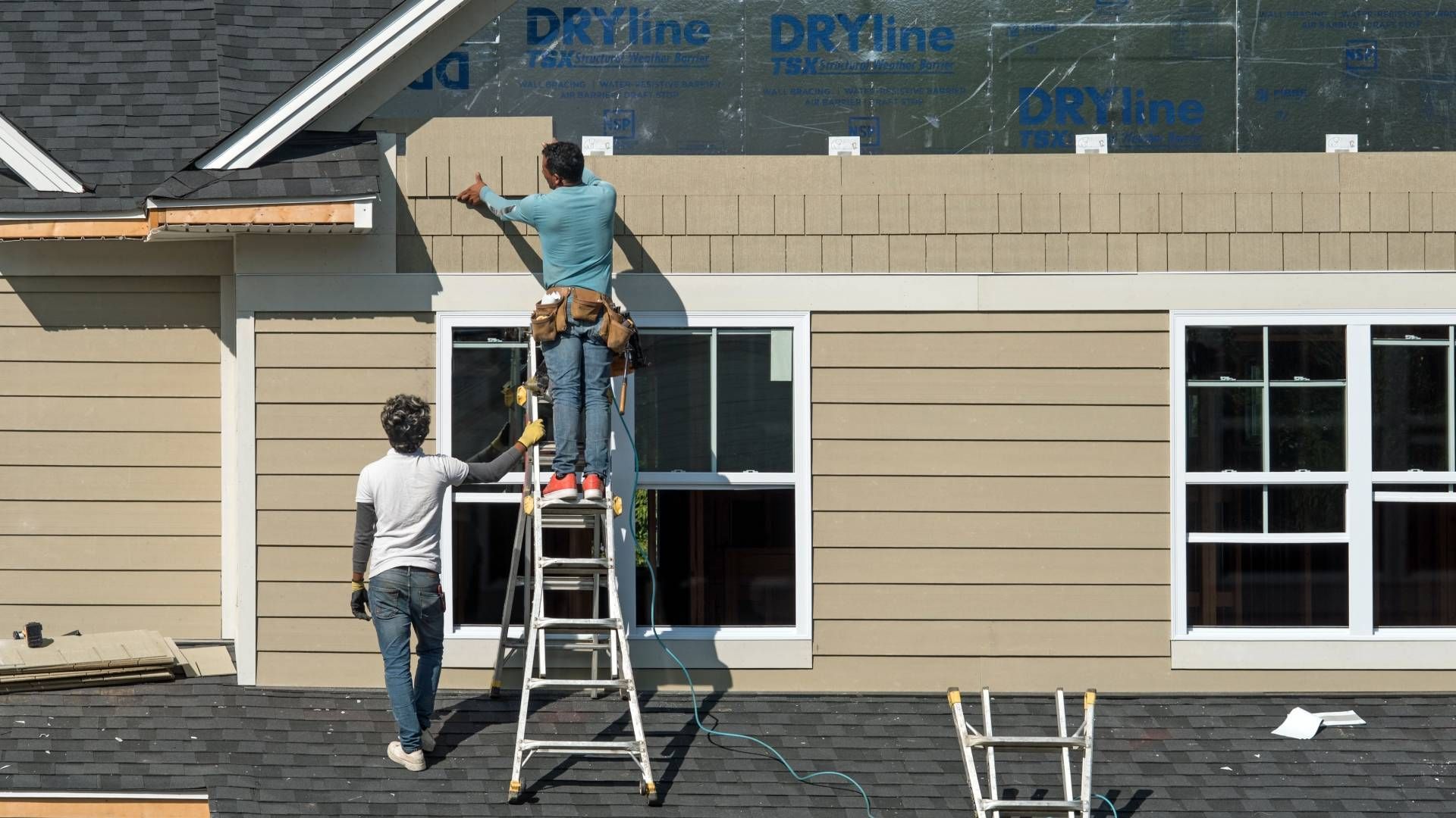 Two people siding a house, one on a ladder installing beige siding.