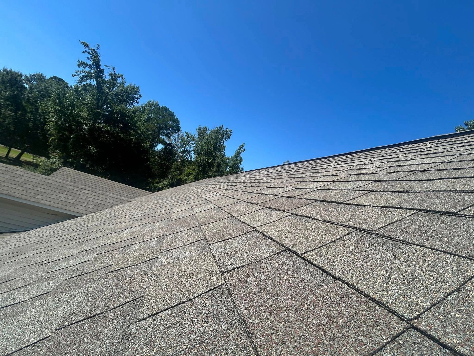 Gray asphalt shingle roof on a sunny day with a bright blue sky and trees in the background.
