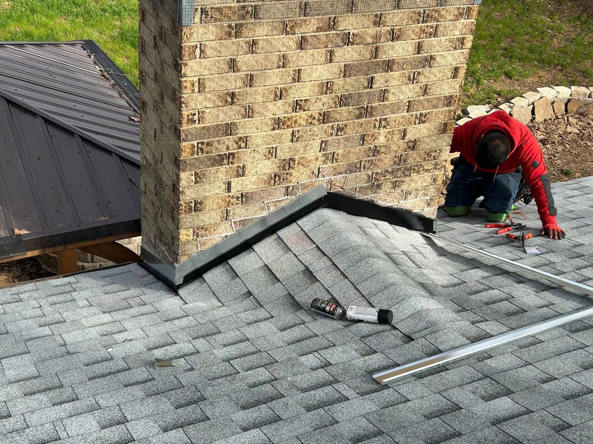 Roofer on a gray shingled roof, flashing around a brick chimney, metal roof in the background.