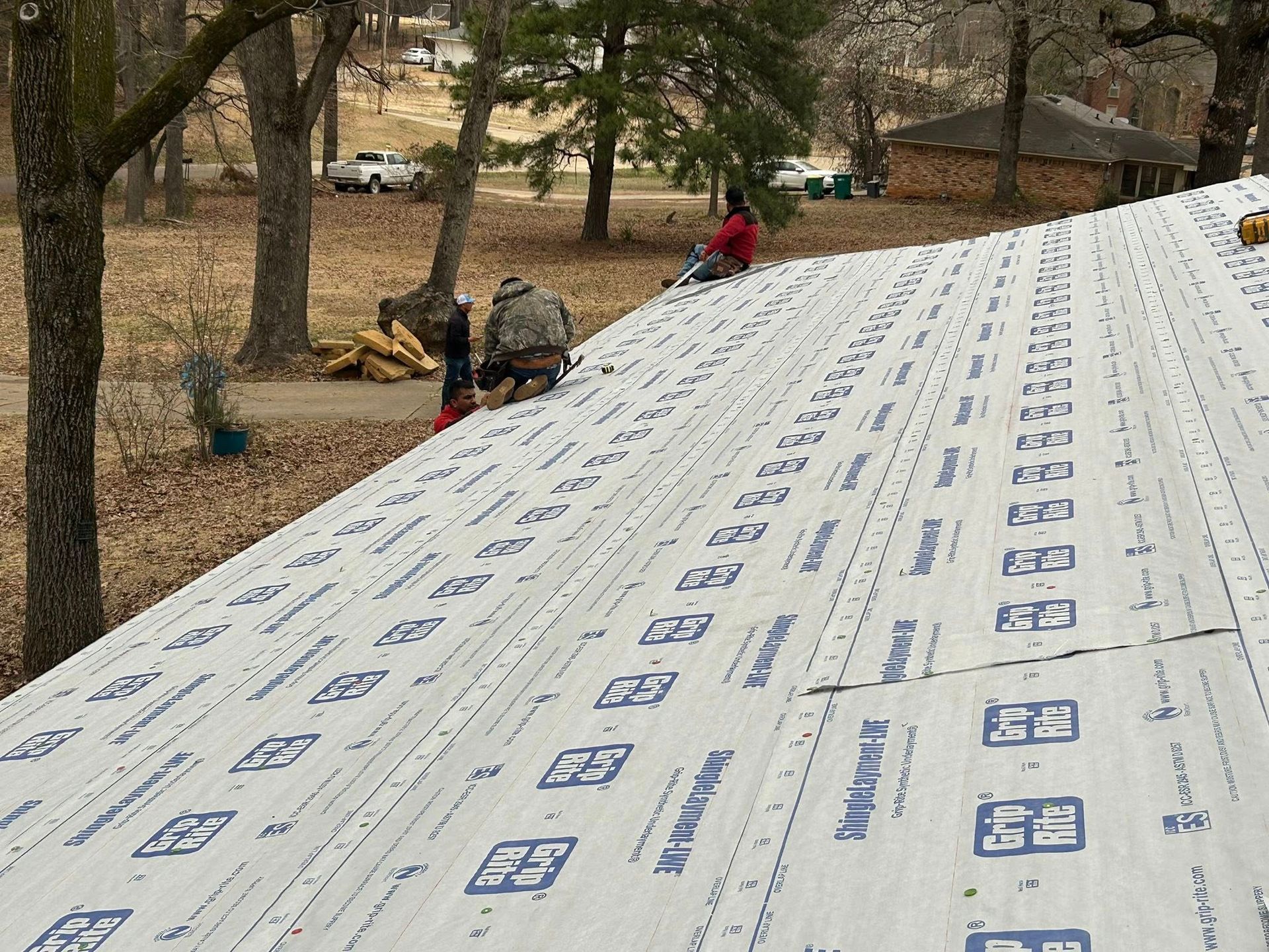 Workers on a roof installing roofing material; trees and houses in the background.